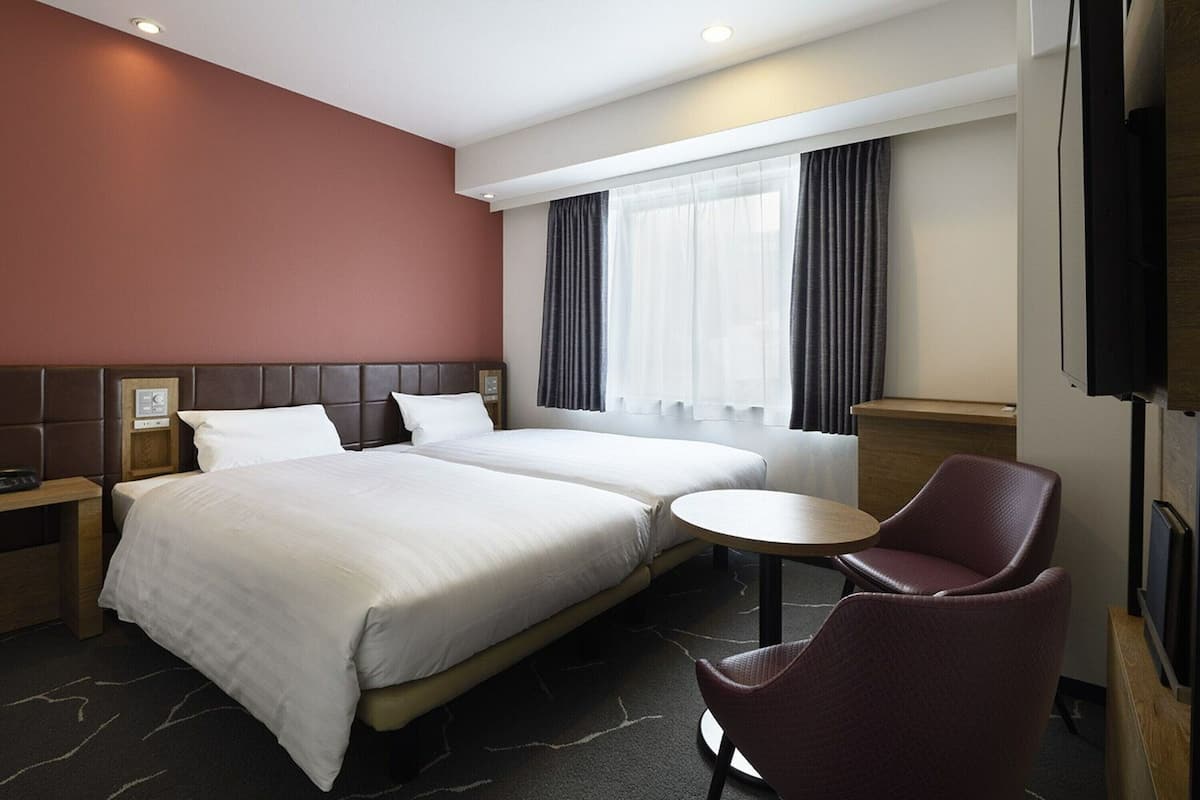 A hotel room with two beds, a small round table, a chair, a TV, and a window with curtains.