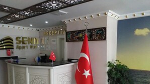 A reception area with a desk, a flag, and a wall with a clock and framed pictures. - Send Apart Otel (Konyaalti)