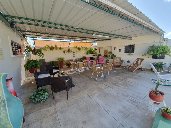 Relax in a serene outdoor space with a metal roof, wicker chairs, and a table, surrounded by potted plants and windows with decorative shutters.