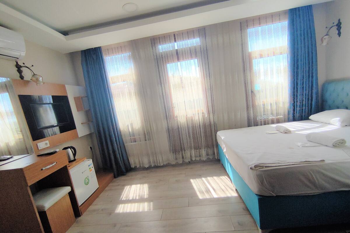 A hotel room with a bed, a desk with a kettle, a television, and a window with curtains.