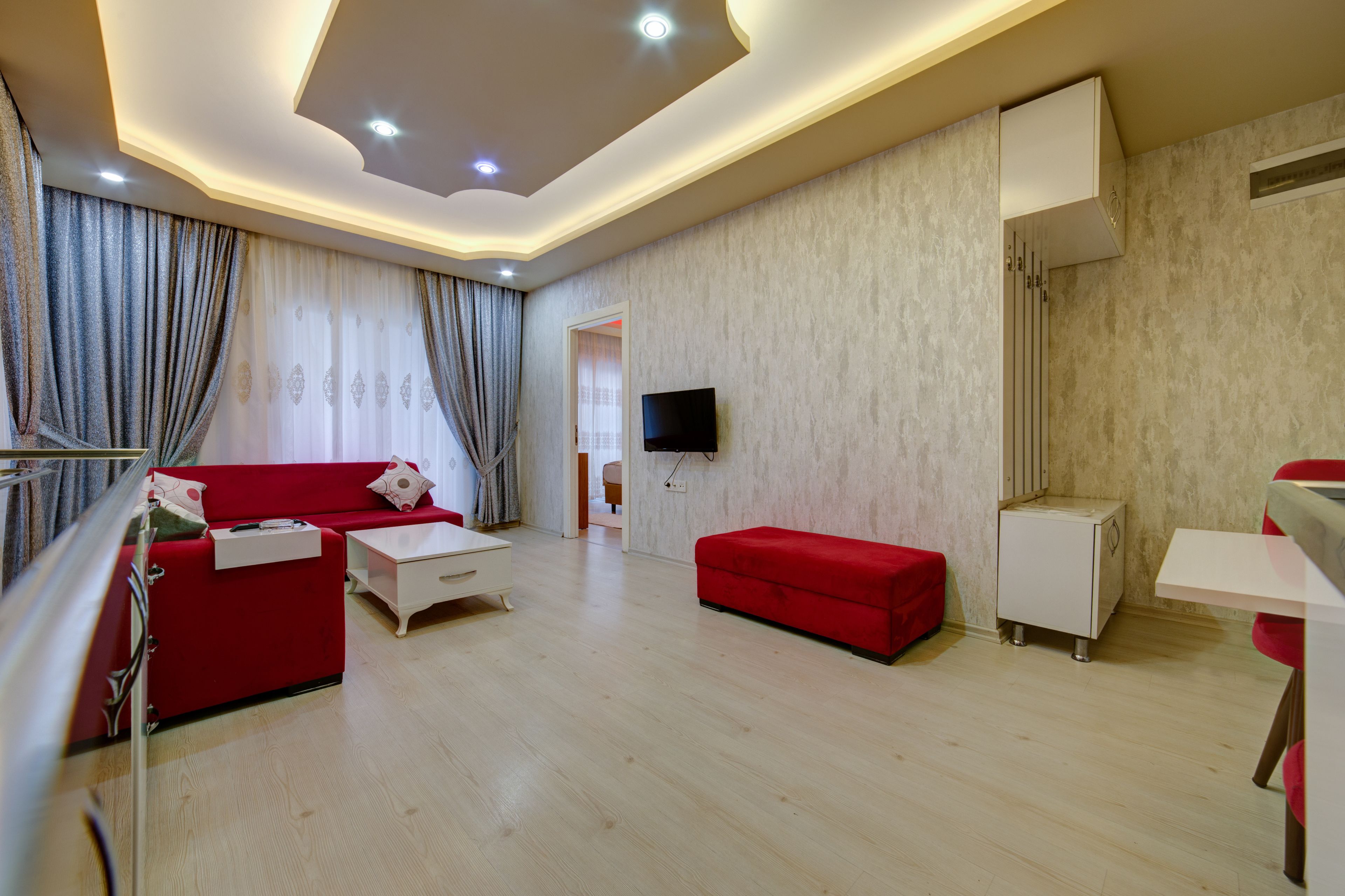 A modern living room with a red sofa, a white coffee table, and a red ottoman. There is a flat-screen TV mounted on the wall, a white cabinet, and a wooden floor.