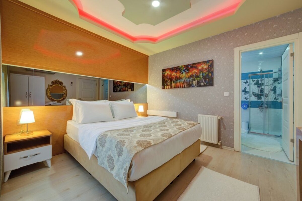 A bedroom with a bed, bedside tables, a nightstand, a mirror, a painting, and a shower area.