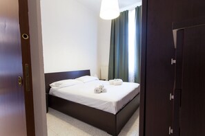 A neatly made bed with white linens and rolled towels, a dark wooden headboard, and a window with green curtains. - Italianway   - Villoresi (Milan)