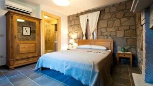A bedroom with a stone wall, a wooden wardrobe, a bed with a blue bedspread, a nightstand with a lamp, and a small table with a green vase. - Idasos Tasodalar (Ayvacik)