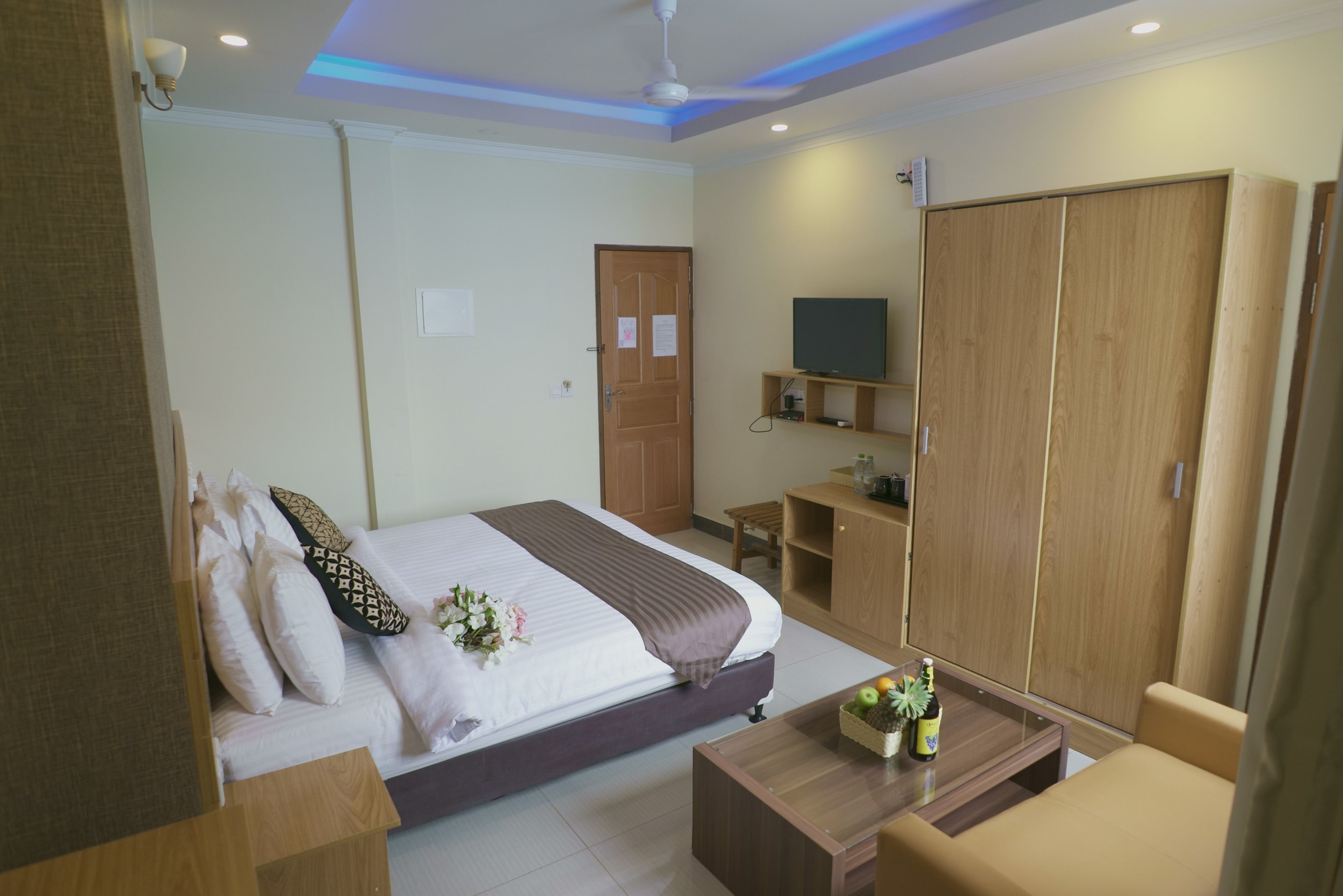 A modern hotel room with a bed, a wooden wardrobe, a TV, and a small table with a plant and a bottle.
