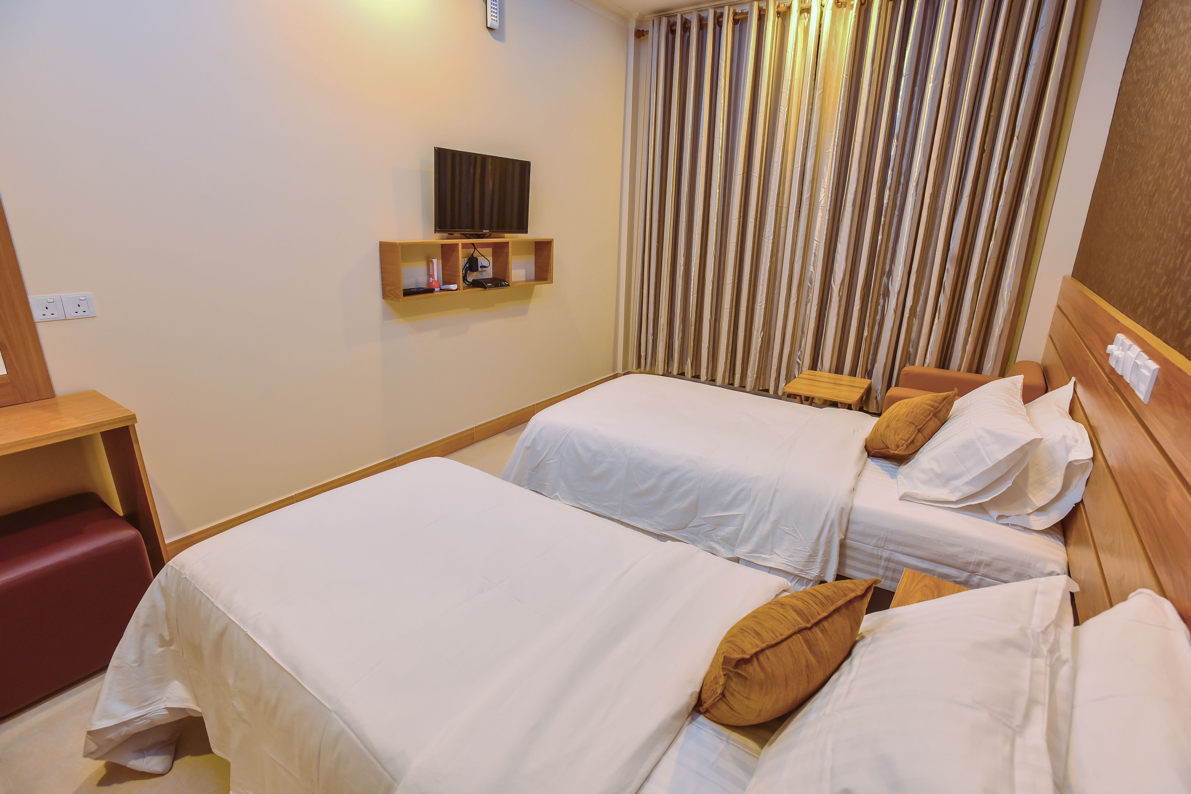 A hotel room with two beds, a TV on a shelf, a red bench, and a curtain.