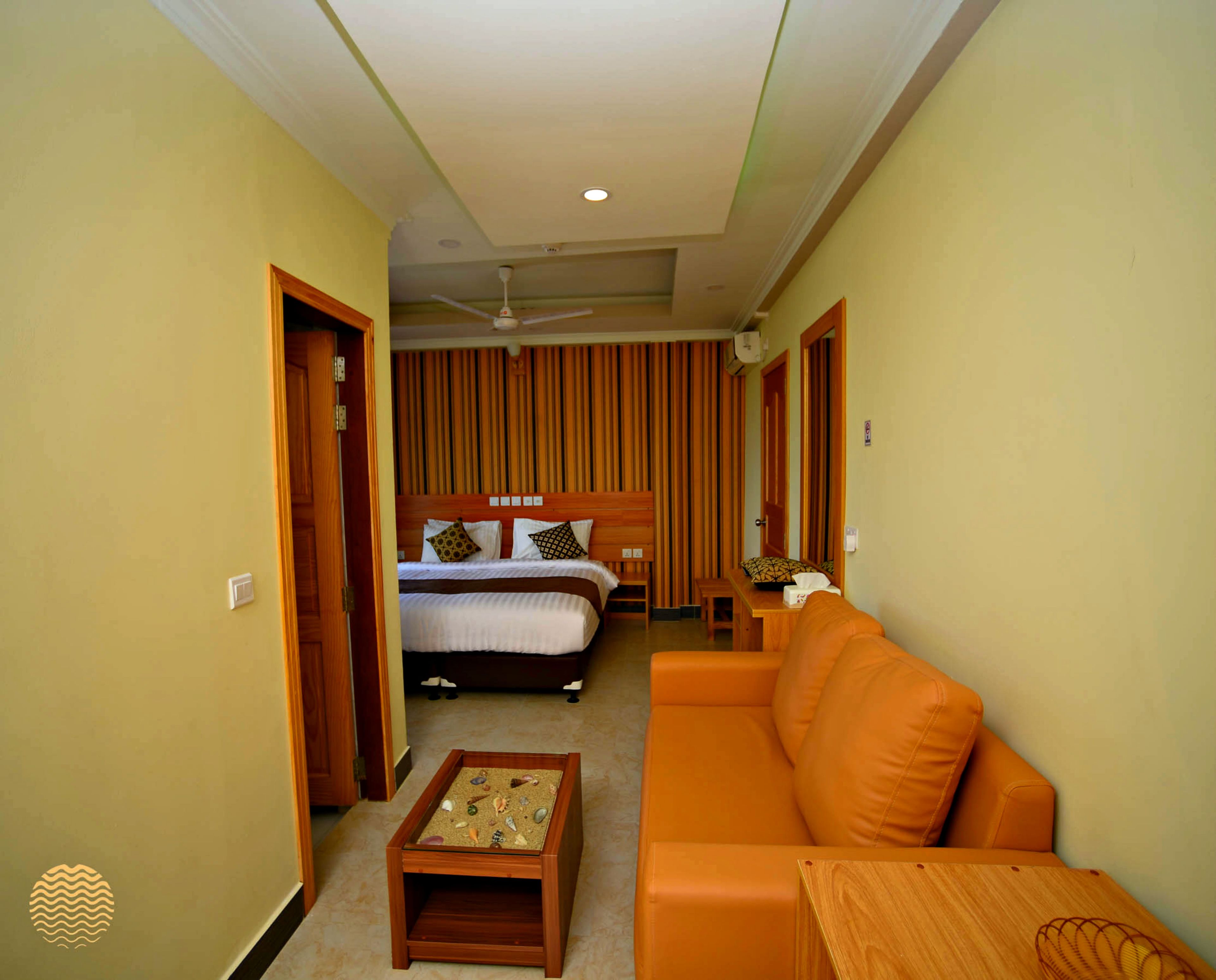 A hotel room with a bed, a wooden nightstand with a decorative item, an orange sofa, and a ceiling fan.