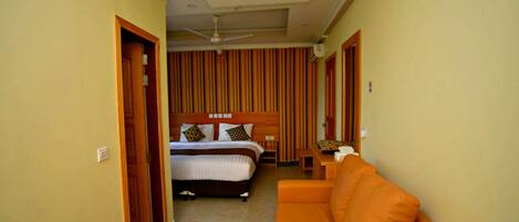 Habitación de hotel con una cama, una mesita de noche de madera con un objeto decorativo, un sofá naranja y un ventilador de techo.
