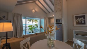 Una sala moderna con vista a la playa, un comedor con mesa y sillas, una cocina con refrigerador y microondas, y un amplio ventanal con cortinas.