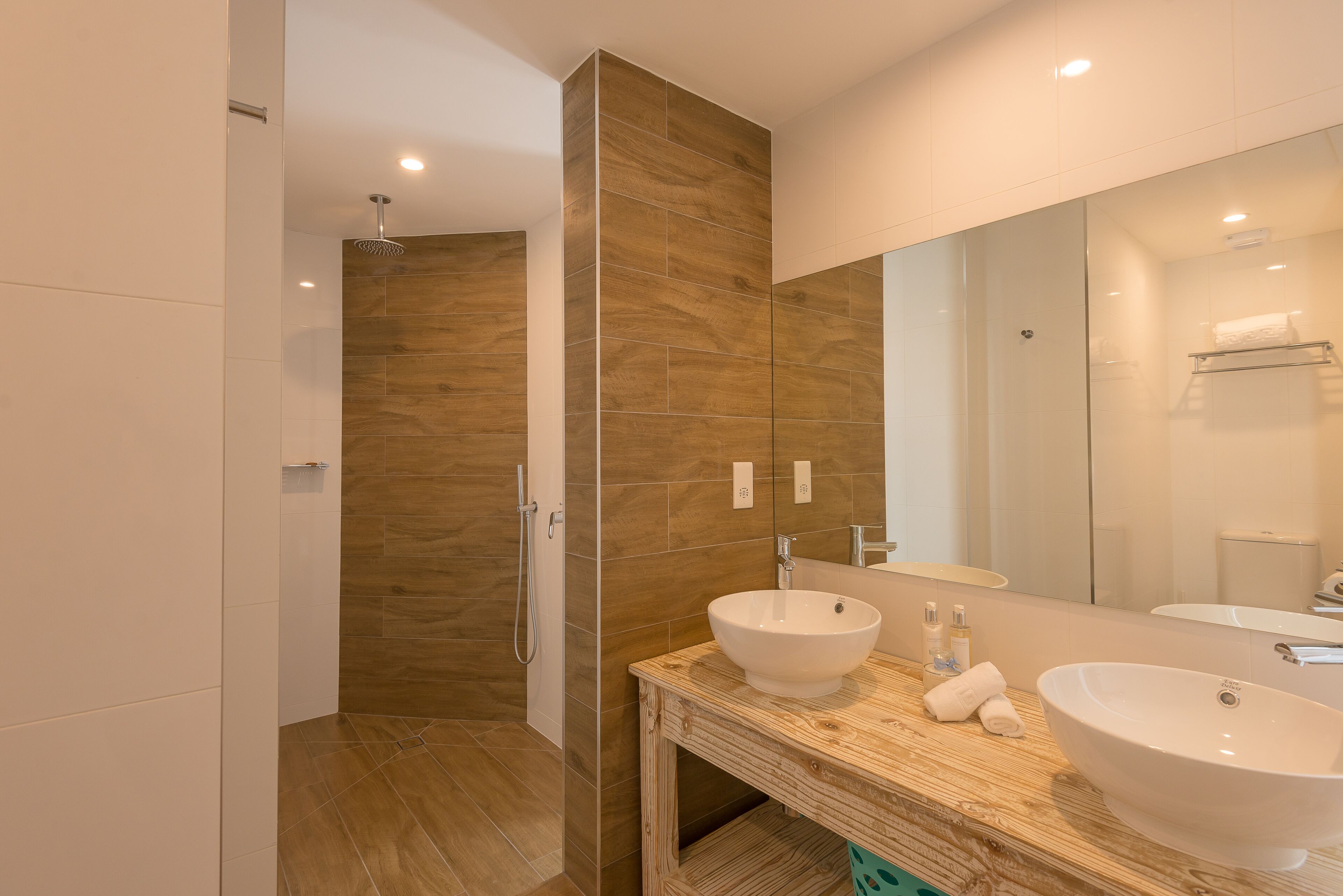 A modern bathroom with a wooden vanity, two sinks, a large mirror, and a walk-in shower.