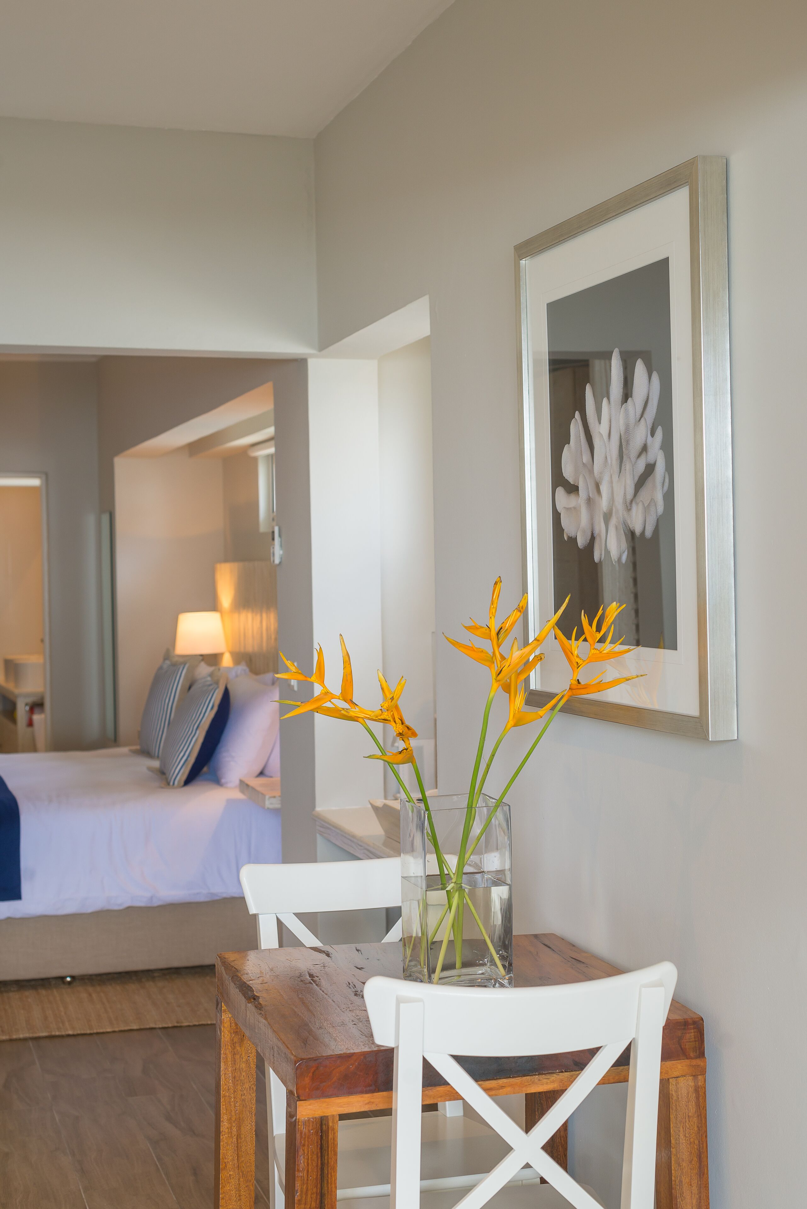 A hotel room with a bed, a bedside table with a vase of flowers, a framed artwork on the wall, and a view into another room.