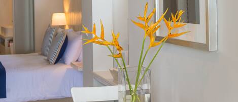 A hotel room with a bed, a bedside table with a vase of flowers, a framed artwork on the wall, and a view into another room.