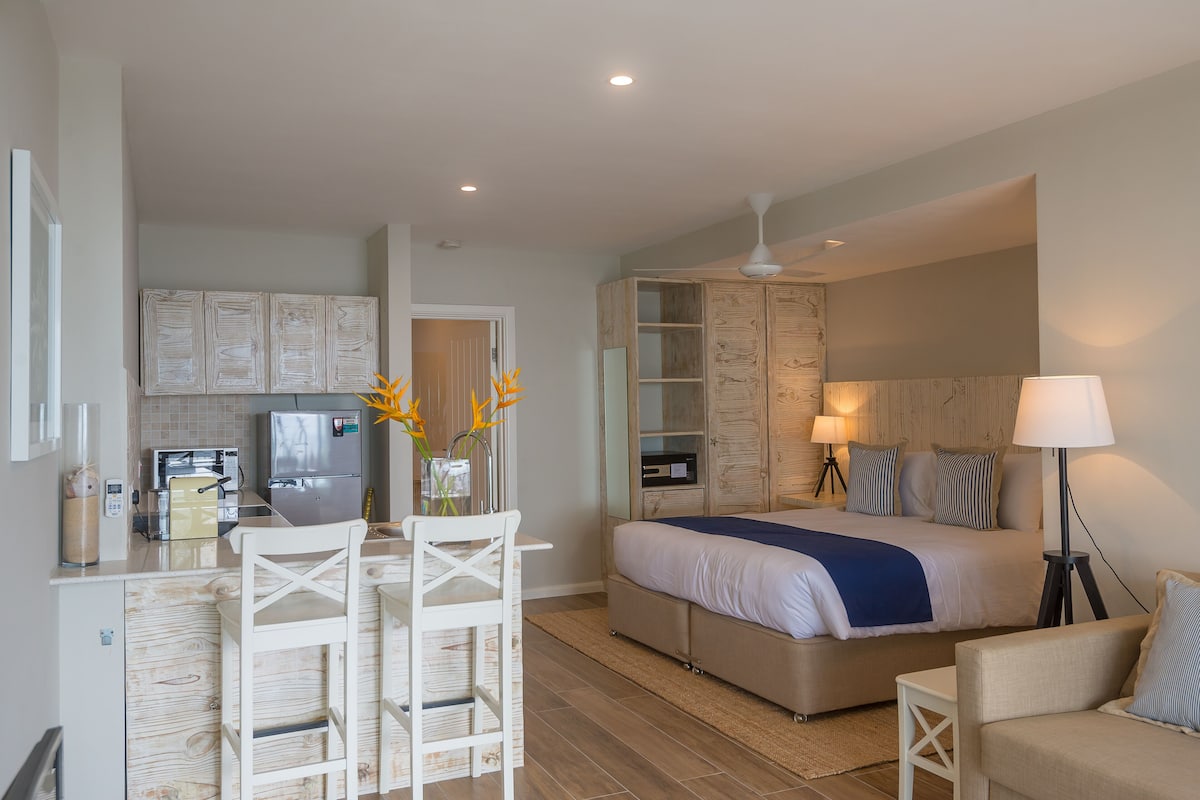 Una habitación de hotel compacta con kitchenette, una cama con manta azul, una mesita de noche con lámpara y un área de descanso con una silla y una pequeña mesa.