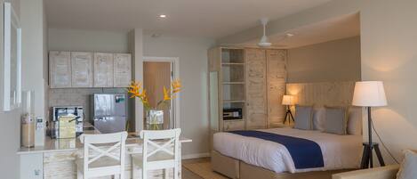 A compact hotel room with a kitchenette, a bed with a blue blanket, a bedside table with a lamp, and a seating area with a chair and a small table.