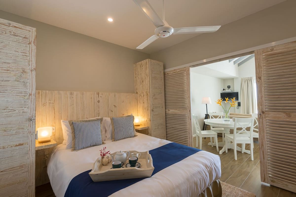 Habitación de hotel con cama, lámparas de noche, ventilador de techo, zona de desayuno con mesa y sillas, y vista al comedor.