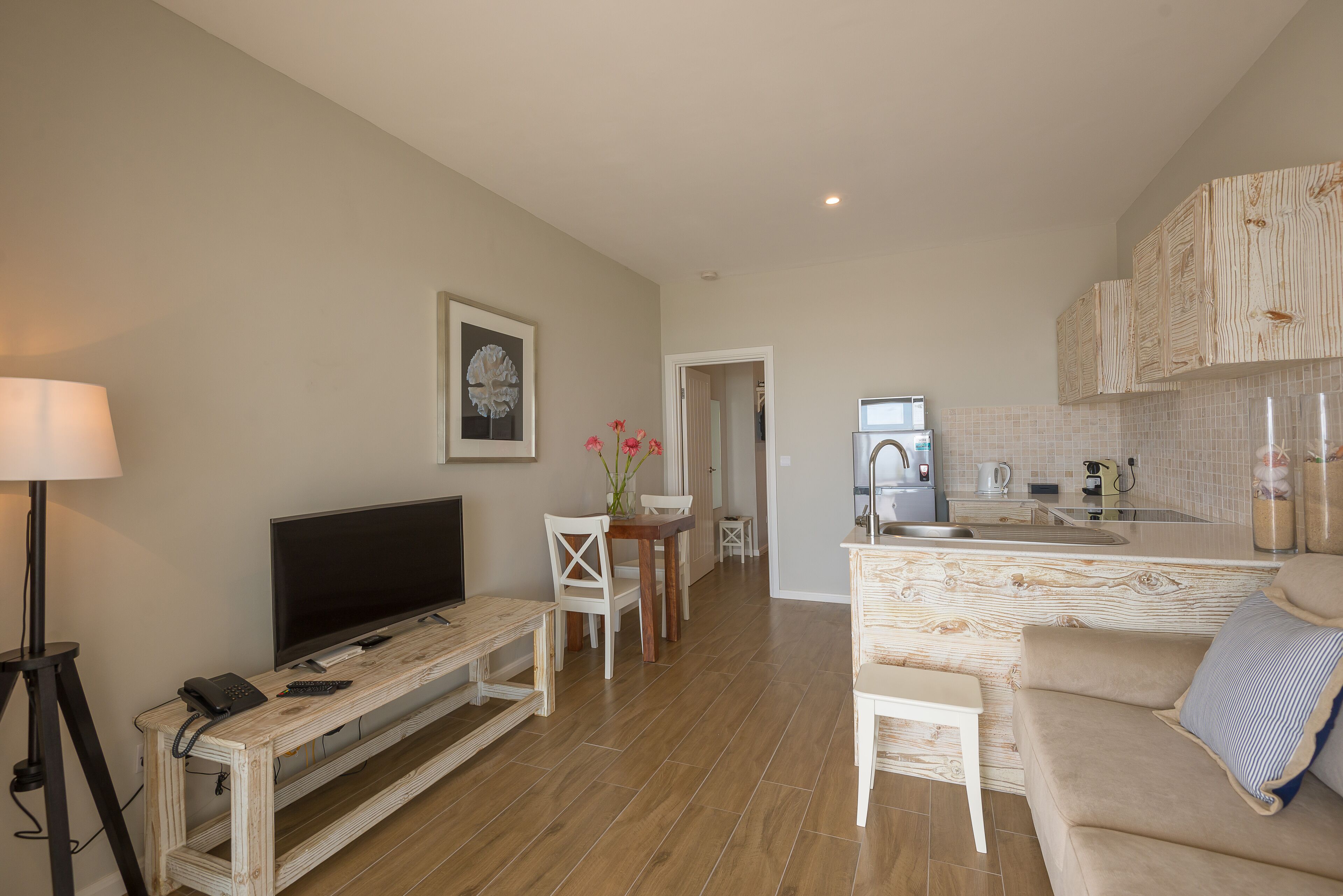 A modern living room with a flat-screen TV, a coffee table, and a sofa. There is a kitchen area with a sink and cabinets.