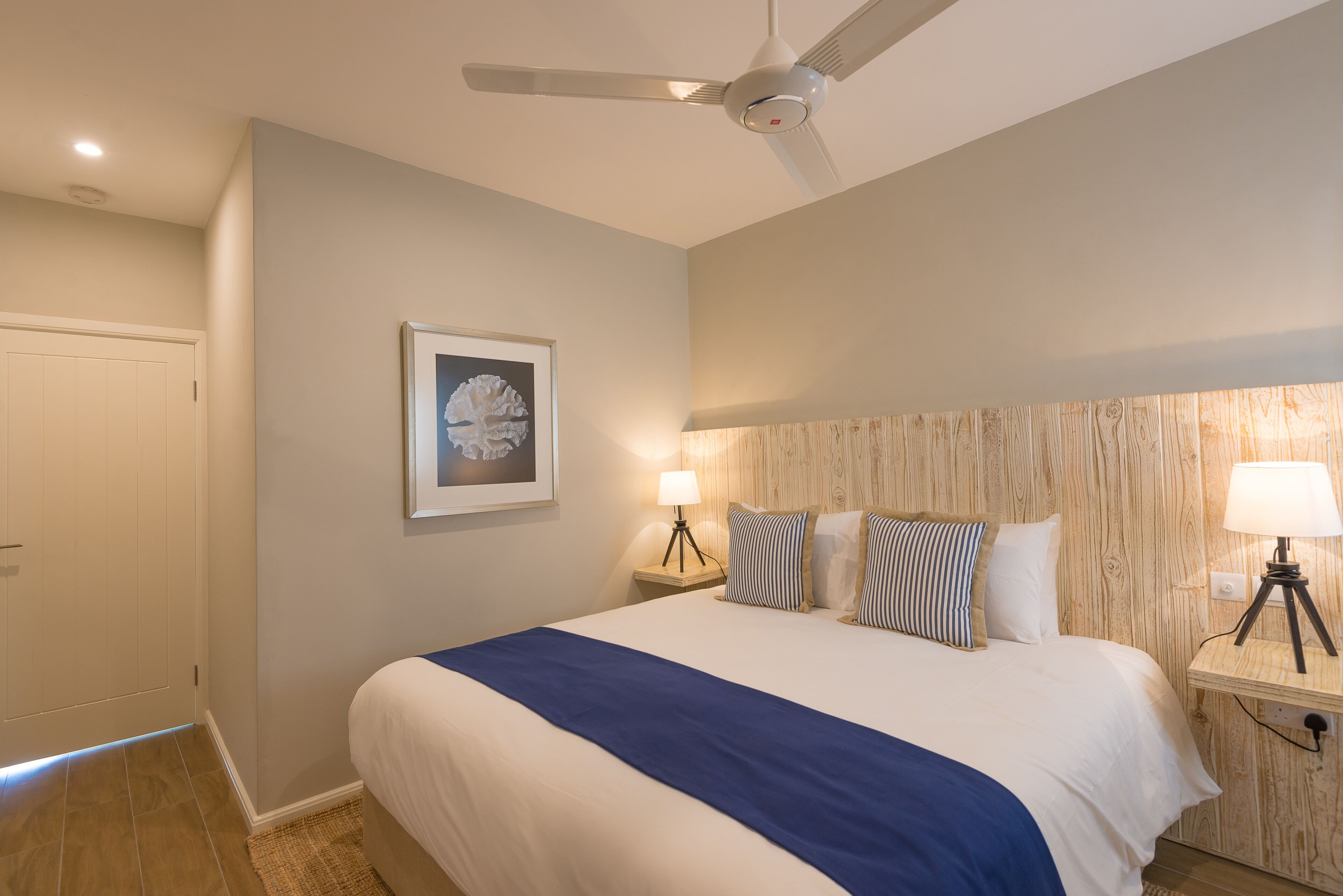 A bedroom with a large bed, two bedside tables with lamps, a ceiling fan, a framed picture on the wall, and a wooden headboard.