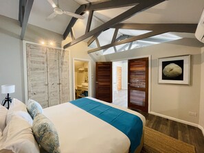 Une chambre avec un lit, un ventilateur de plafond, des poutres en bois, une salle de bain visible à travers une porte ouverte et un tableau encadré au mur.