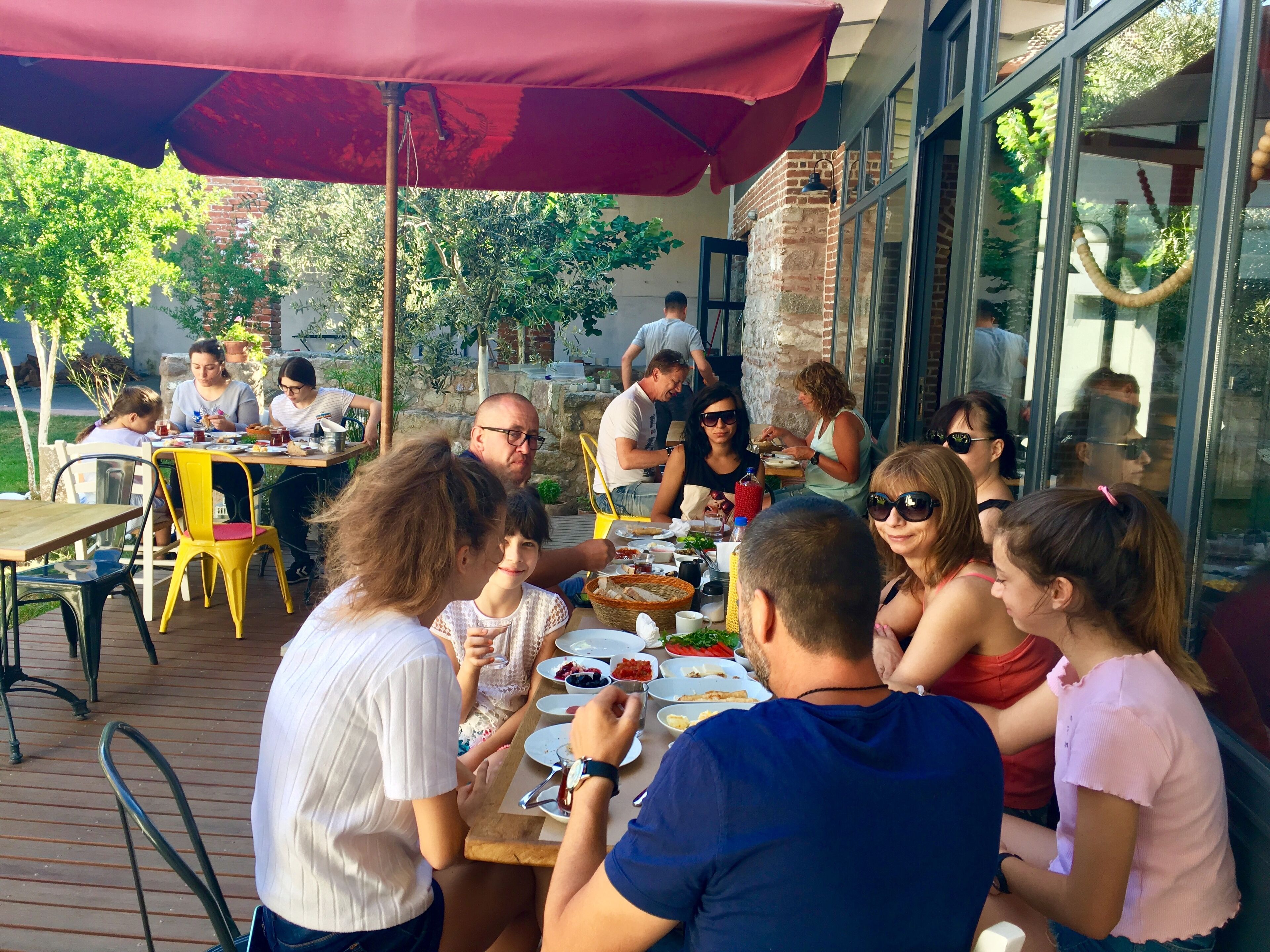 A group of people dining outdoors at a table with a red umbrella, surrounded by brick walls and large windows.