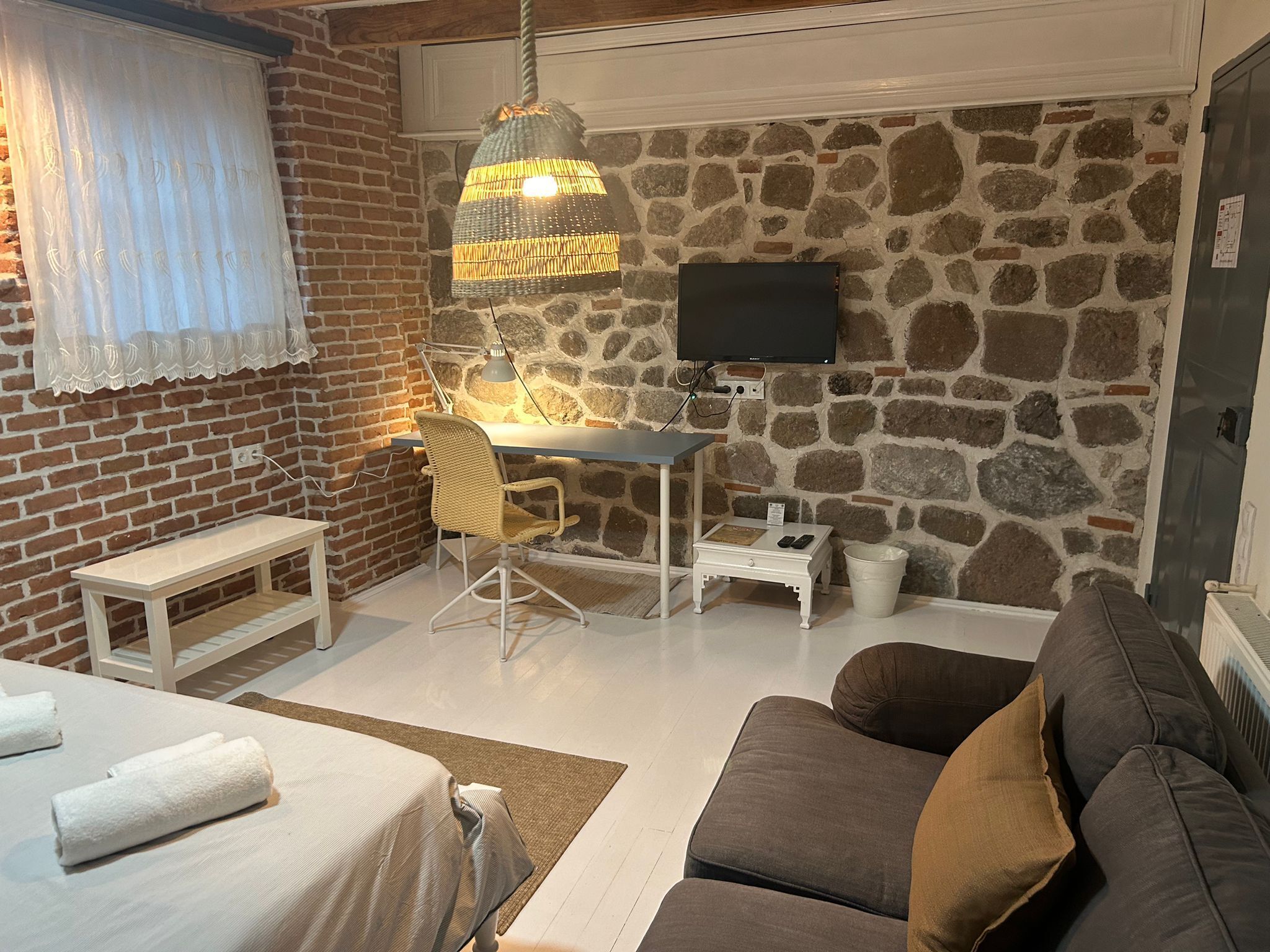 Una habitación acogedora con pared de ladrillo y pared de piedra, una cama con ropa blanca, un sofá marrón, una mesita pequeña, una silla, un escritorio con lámpara y un televisor montado en la pared de piedra.