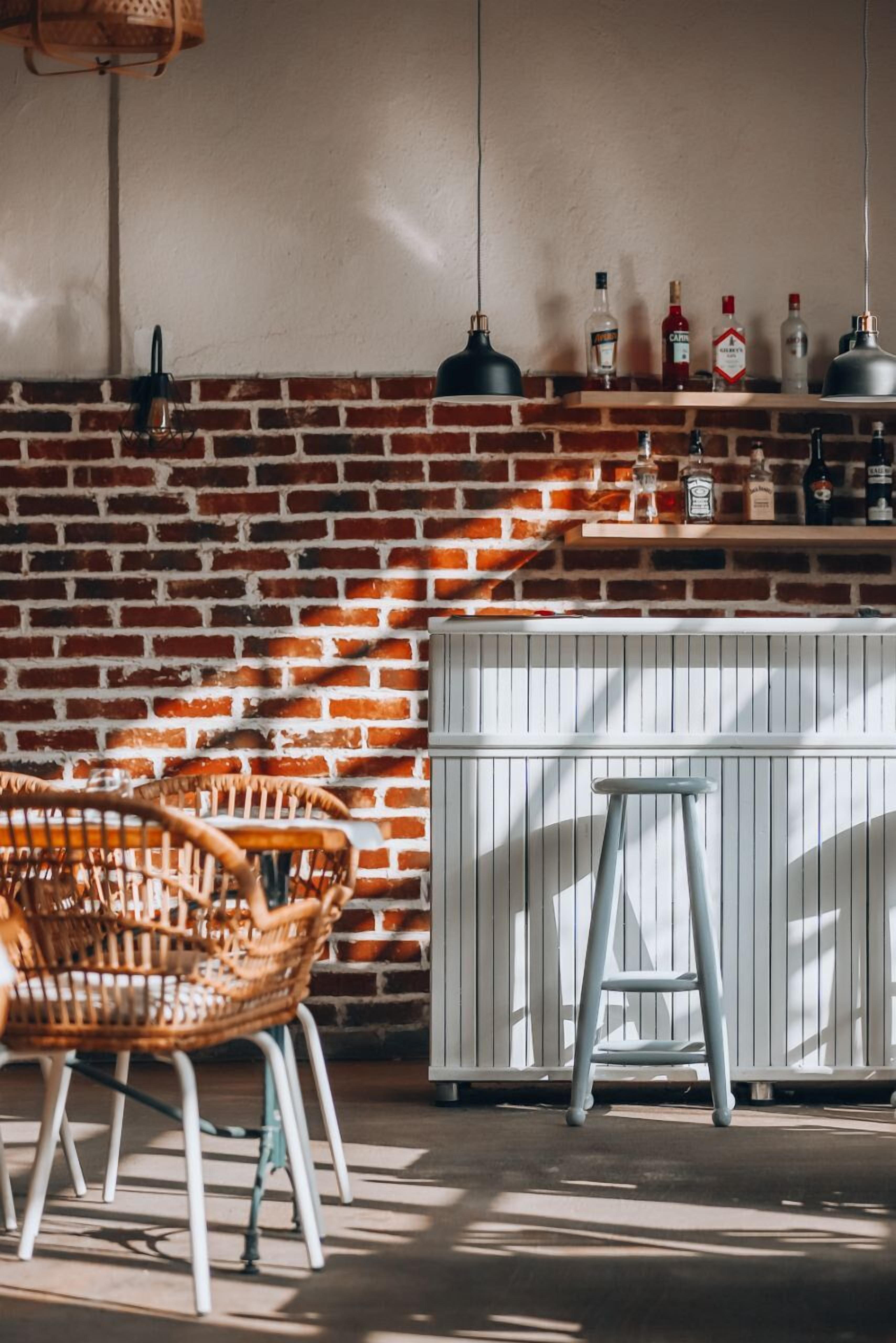 A bar area with a brick wall, wicker chairs, a stool, and a shelf with bottles.