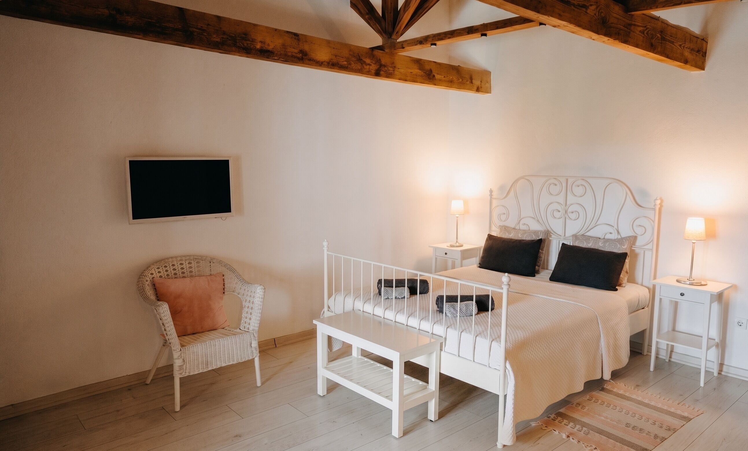 Een slaapkamer met een bed, nachtkastjes, een stoel, een televisie en zichtbare houten balken.