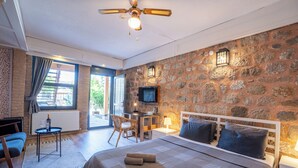 A bedroom with a stone wall, a bed with pillows, a desk with a chair, a television, and a ceiling fan. - Attalos Suites Hotel (Bergama)