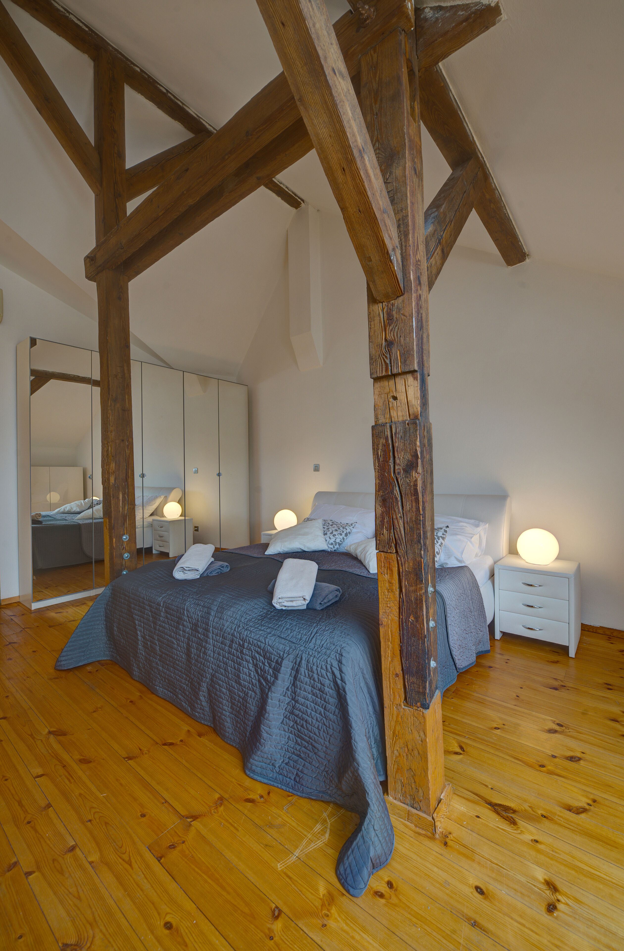 A bedroom with a wooden ceiling, a large bed with a blue blanket, two bedside tables with lamps, and a wardrobe with a mirror.