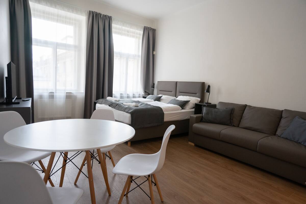 A modern hotel room with a bed, a sofa, a dining table with chairs, and a TV.
