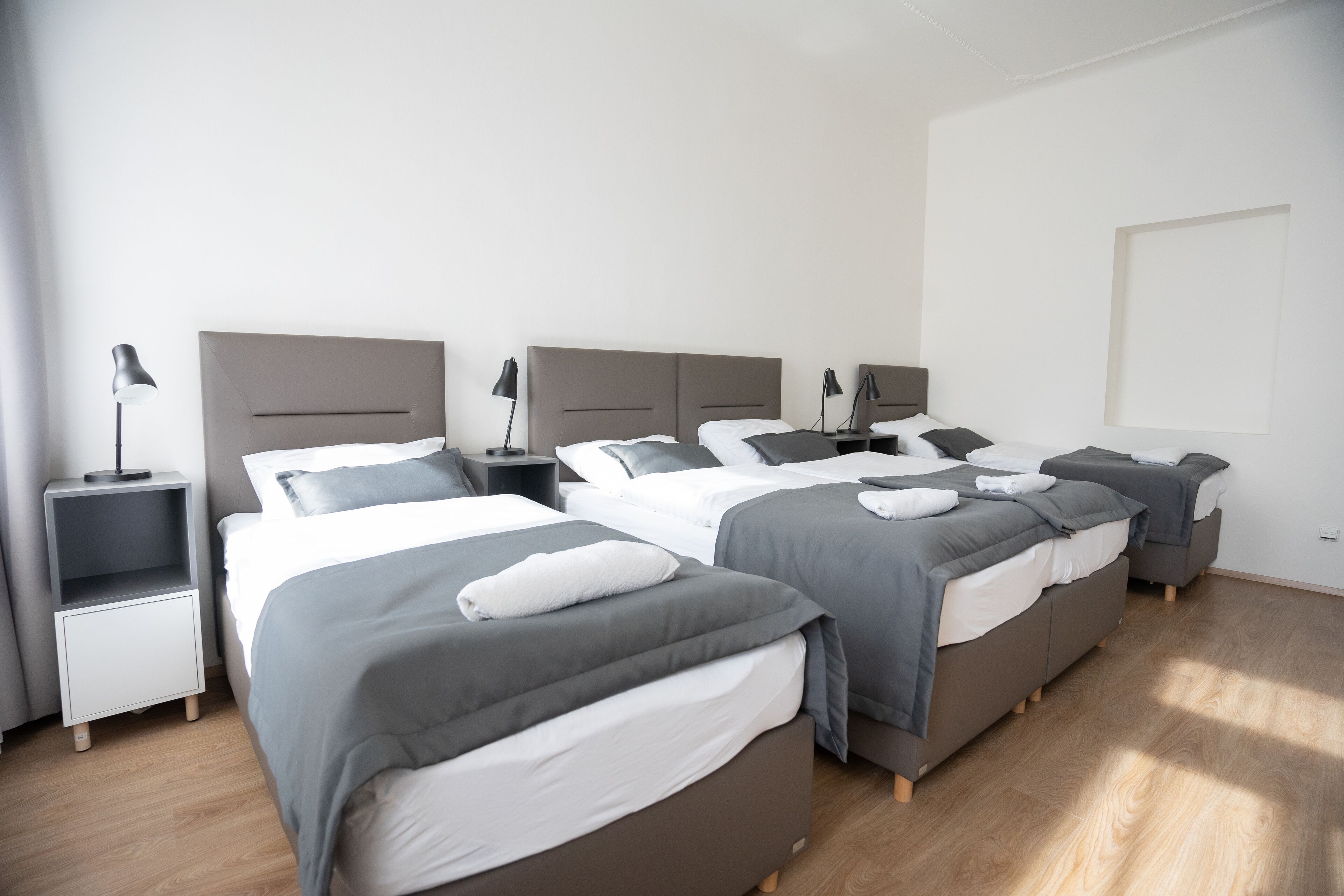 A hotel room with three beds, each with grey bedding and white pillows. There are bedside tables with lamps and a small shelf. The room has wooden flooring and white walls.