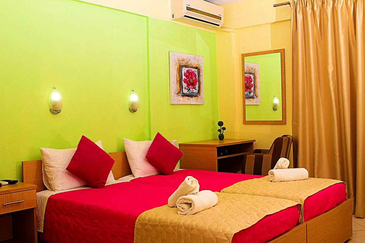 Een hotelkamer met twee bedden, elk voorzien van rood en beige beddengoed, een houten bureau, een stoel en twee wandgemonteerde airconditioners.
