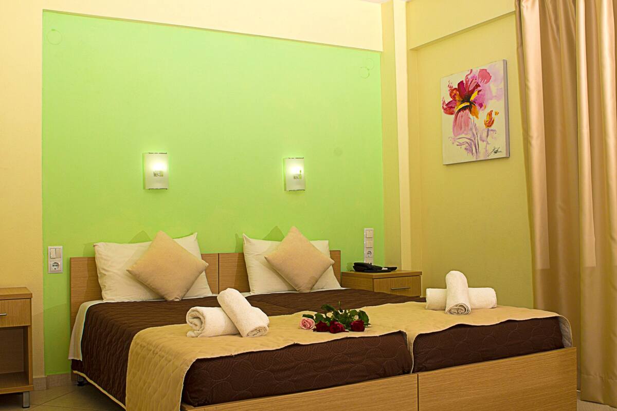 Hotelkamer met een groene muur, twee nachtkastjes, een bed met een bruine hoofdeinde, beige kussens en een schilderij van een bloem.