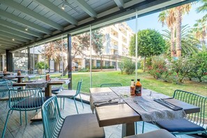 An outdoor dining area with tables and chairs, a glass wall, and a view of a building and greenery. - TUI FAMILY LIFE Islantilla (Isla Cristina)