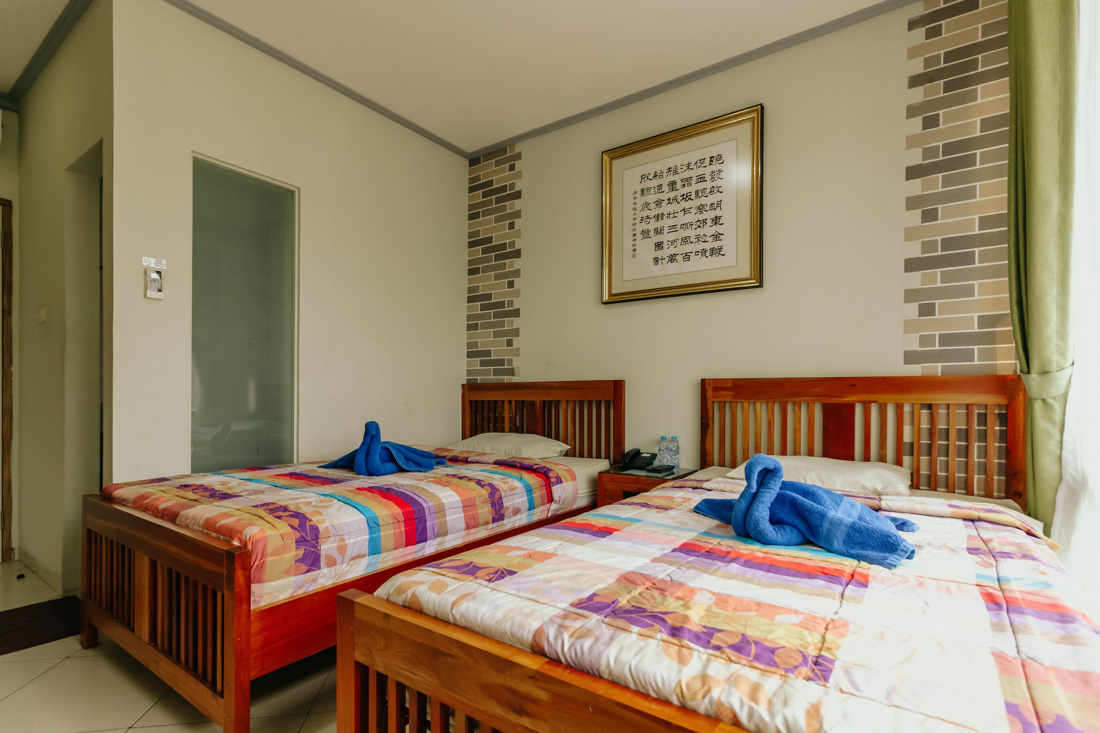 Een hotelkamer met twee houten bedden, elk voorzien van kleurrijke beddengoed en blauwe handdoeken. Er hangt een tekst in een lijst aan de muur en links is een deur.