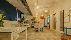 A modern interior with a dining area, chairs, tables, potted plants, and a view of the city at night. - Urbanest Inn House TB Simatupang (Jakarta)