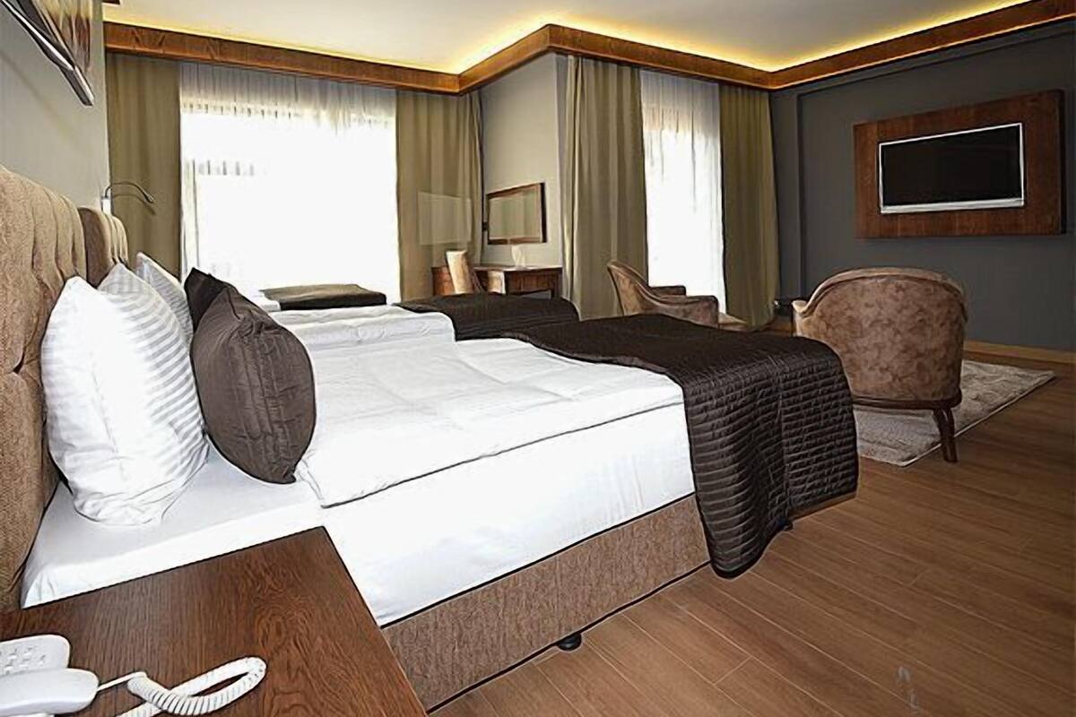 A hotel room with a large bed, a bedside table with a phone, a chair, a desk with a lamp, and a TV mounted on the wall.