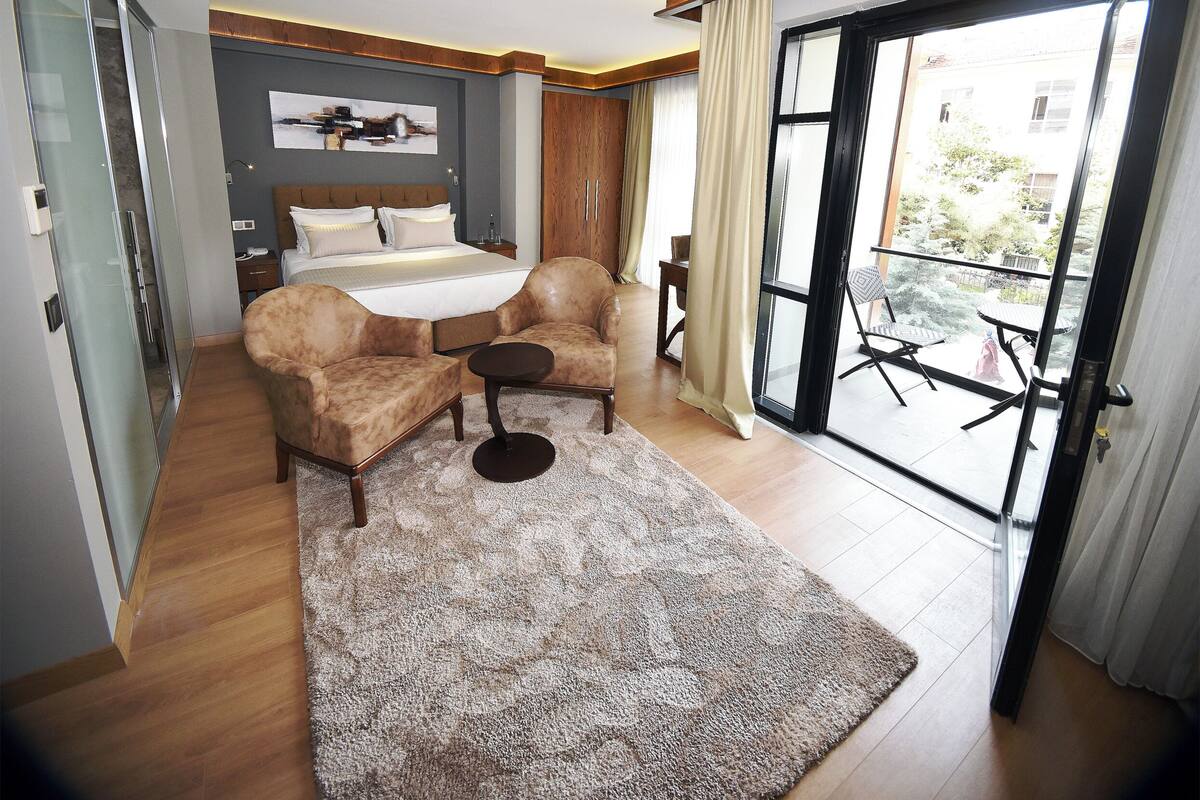 A modern bedroom with a bed, two armchairs, a small table, and a balcony with a view.