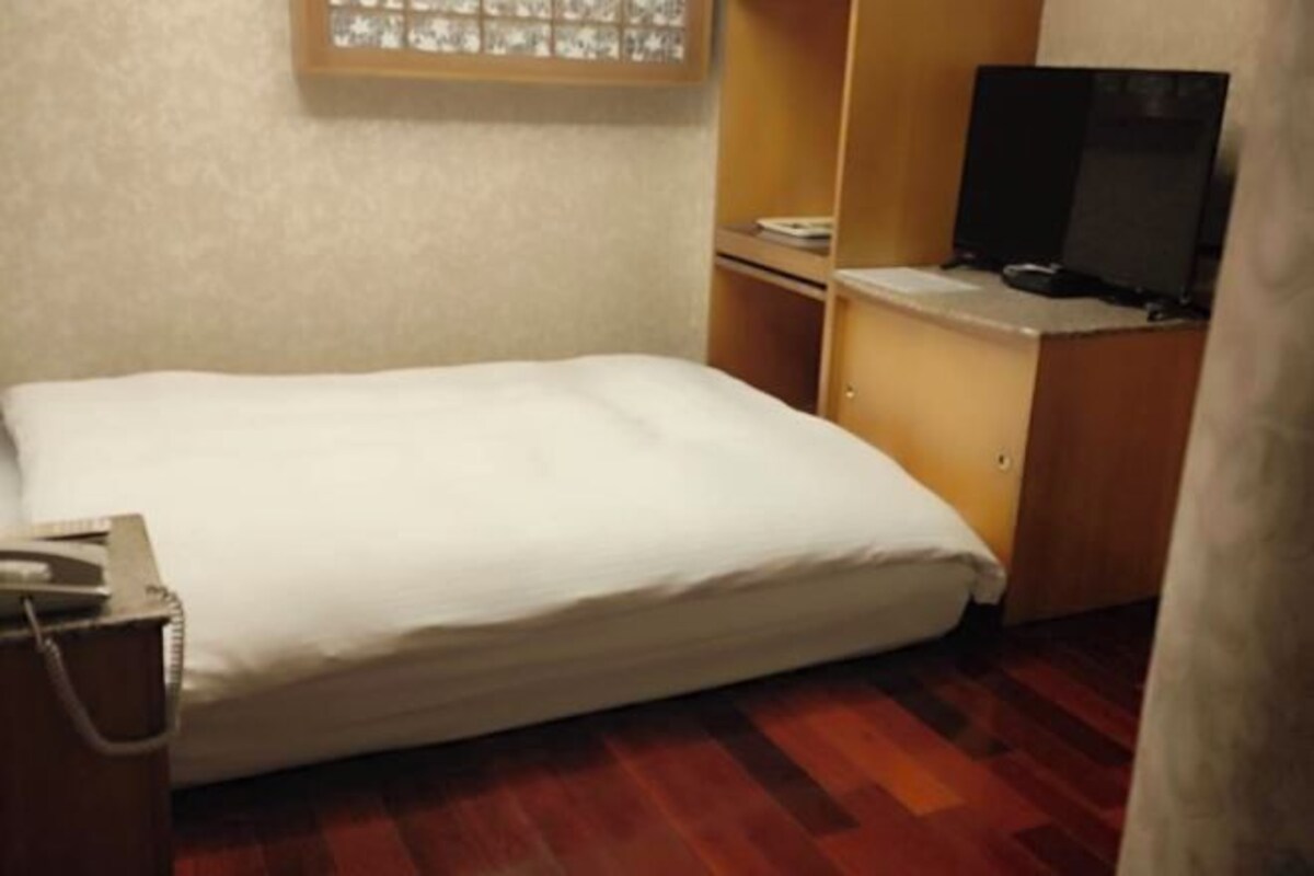 A small, neatly made room with a single bed, a wooden desk, a television, and a window with blinds.