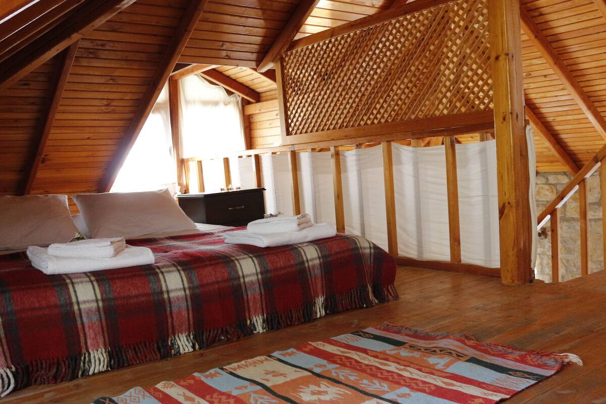 Un dormitorio acogedor en el  ático, con techo de madera, colcha de cuadros y alfombra estampada.