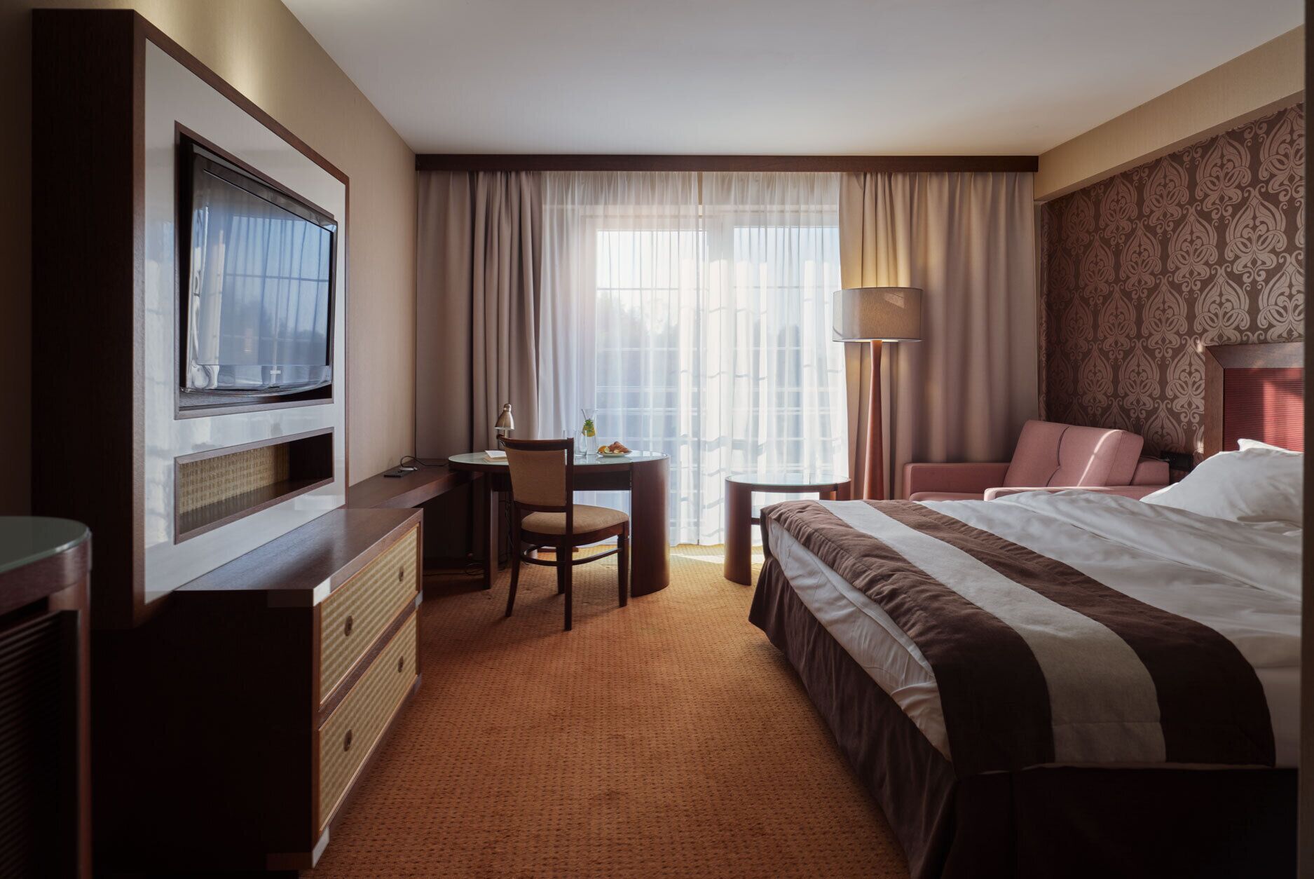A hotel room with a bed, a desk with a chair, a television, and a window with curtains.