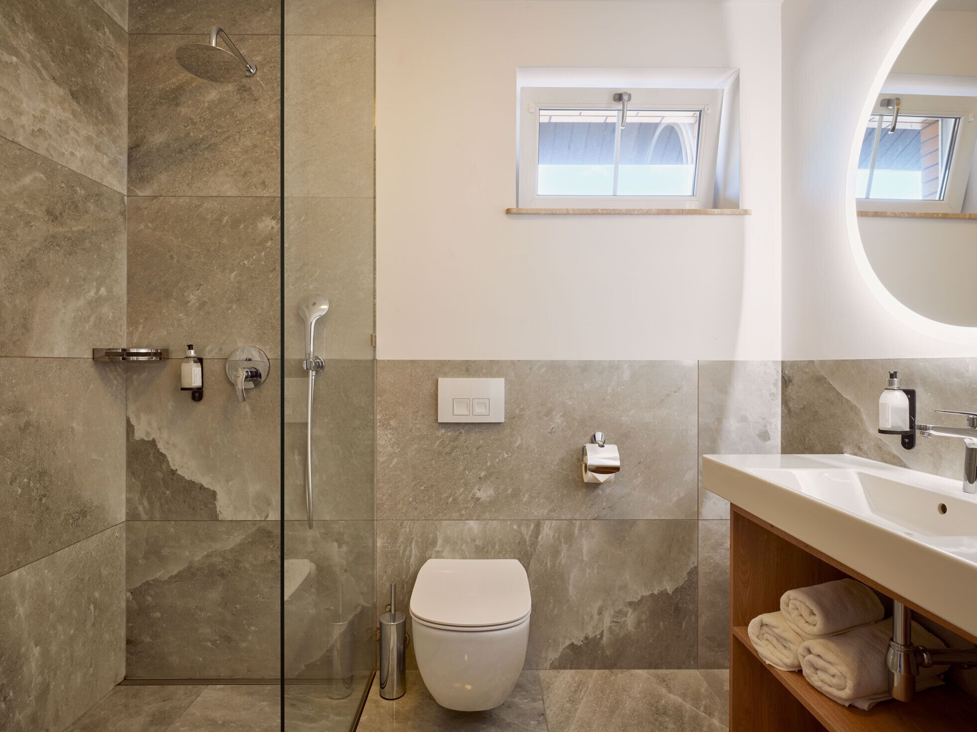 Een moderne badkamer met een douchecabine van glas, een toilet, een wastafel met spiegel en een raam boven de wastafel.