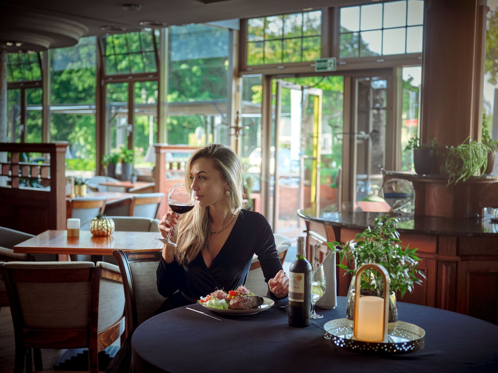 Eine Person sitzt an einem Tisch mit einem Teller Essen, einer Weinflasche und einer Kerze. Im Hintergrund sind große Fenster mit Blick auf eine grüne Landschaft zu sehen.