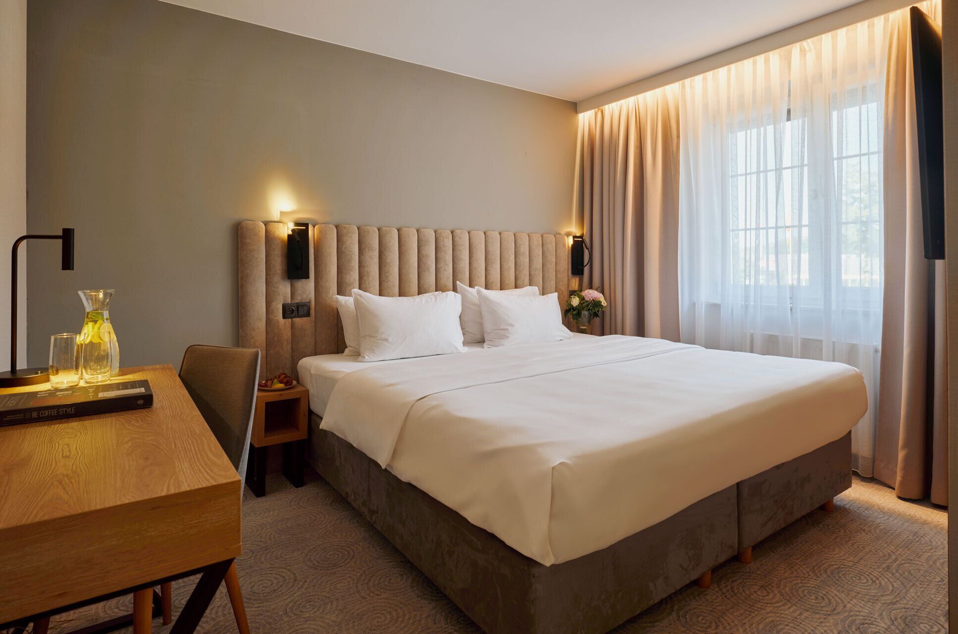Habitación de hotel con una cama grande, una mesita de noche con lámpara, una silla y vistas al exterior a través de cortinas translúcidas.