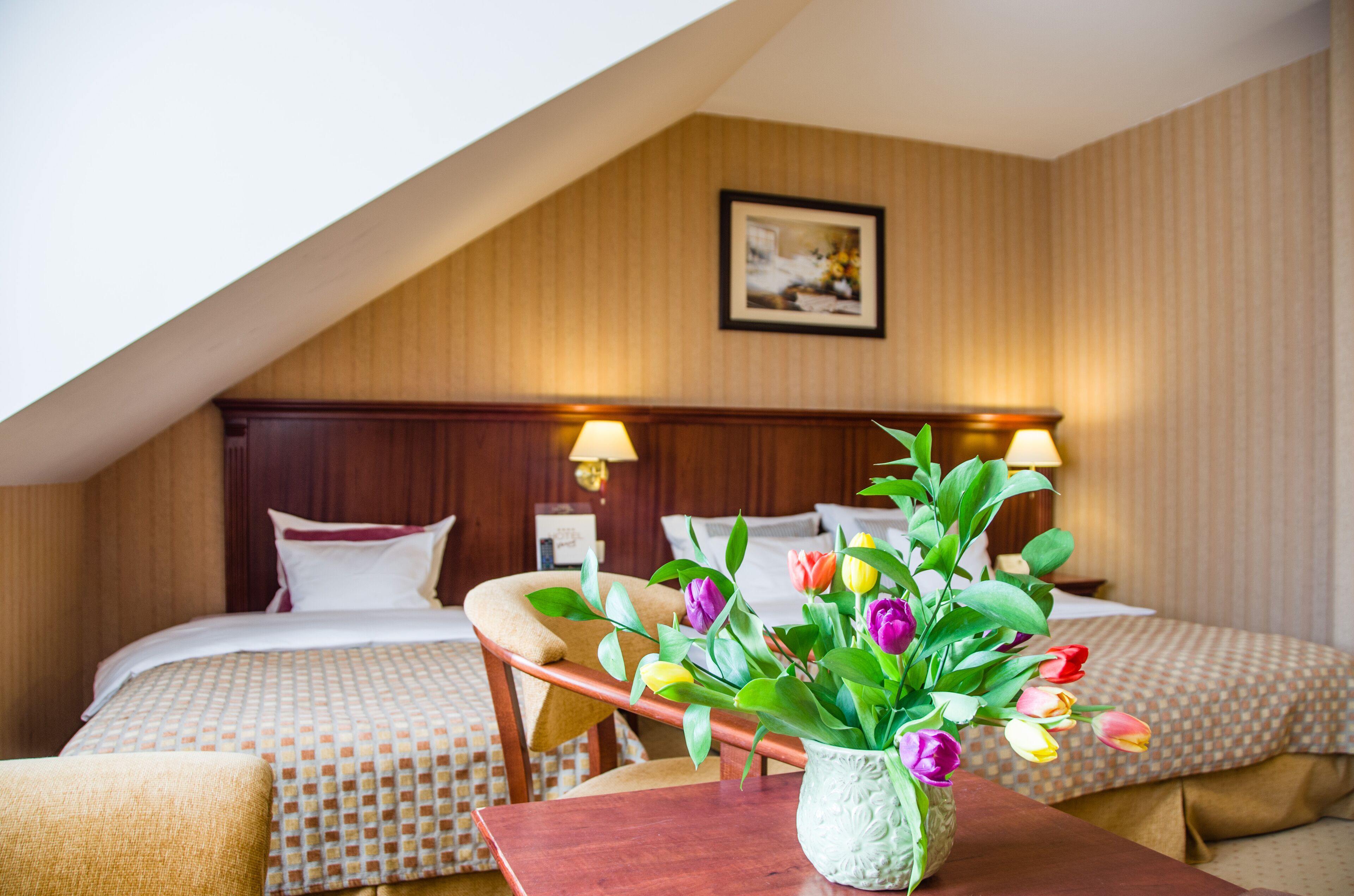 A hotel room with two beds, a wooden headboard, a nightstand with a lamp, a table with a vase of tulips, and a painting on the wall.