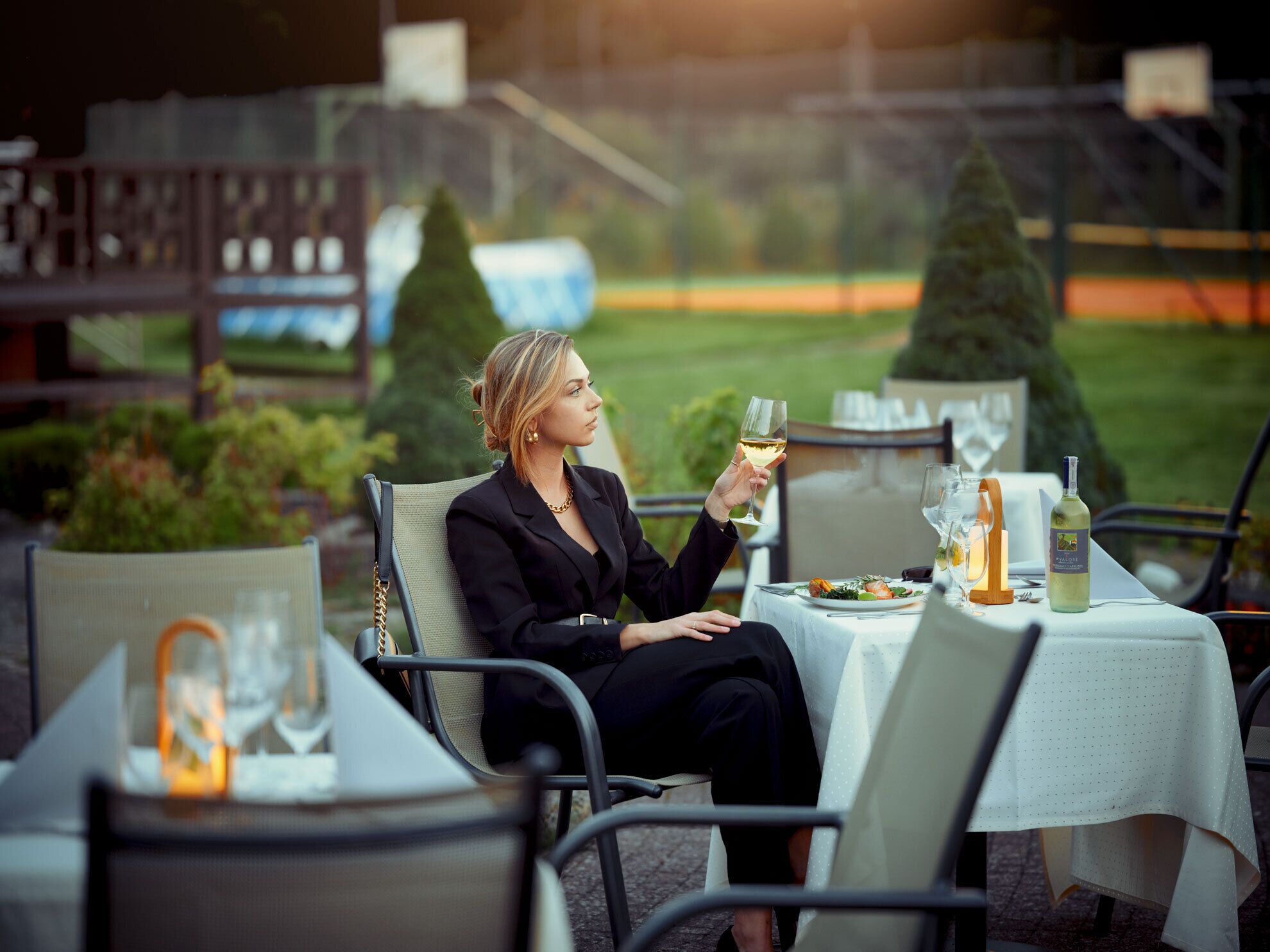 A person seated at an outdoor table with a meal and a glass of white wine.