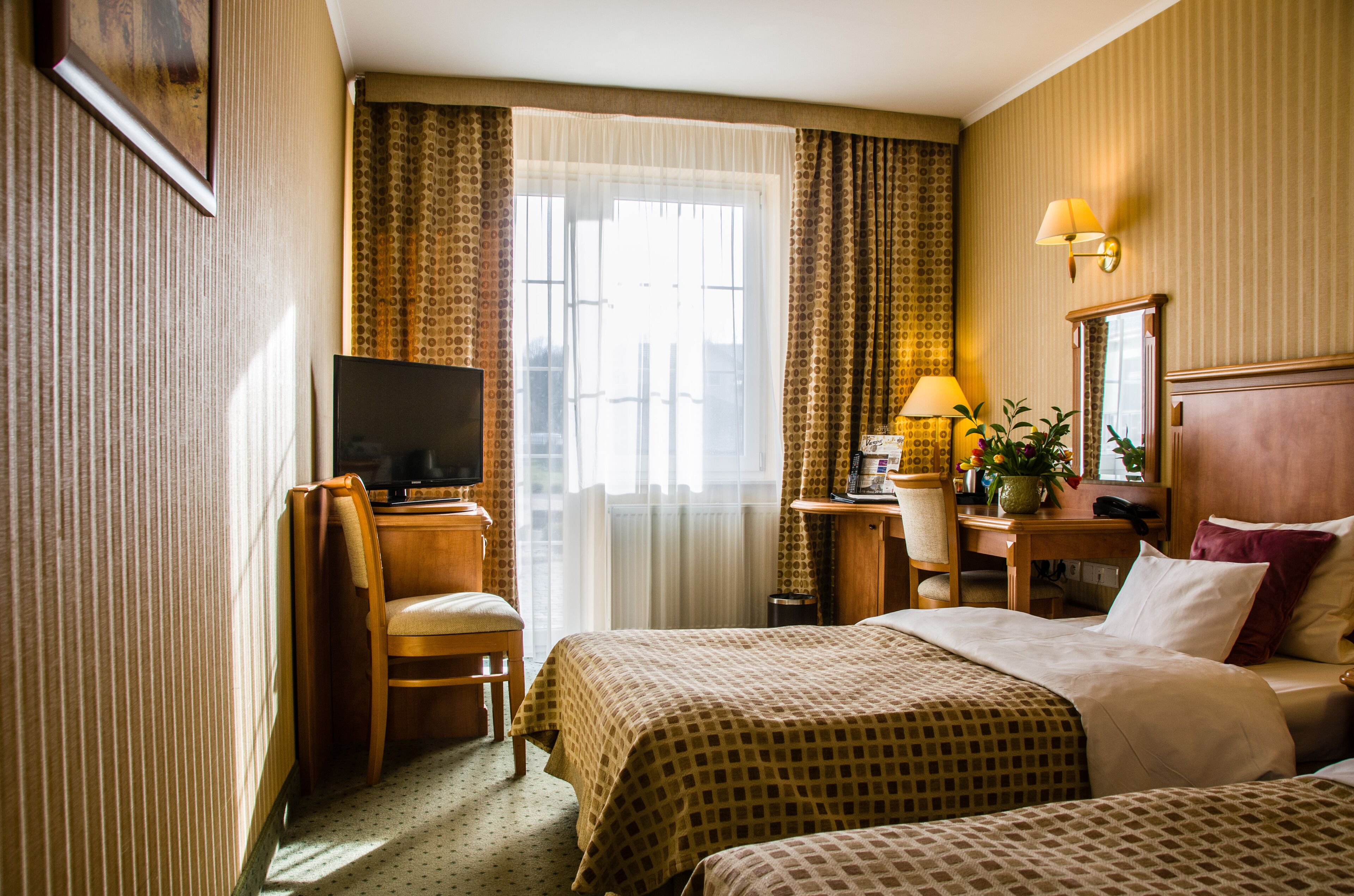 Une chambre d’hôtel avec deux lits, un bureau avec une chaise, une télévision, une lampe et une fenêtre avec des rideaux.