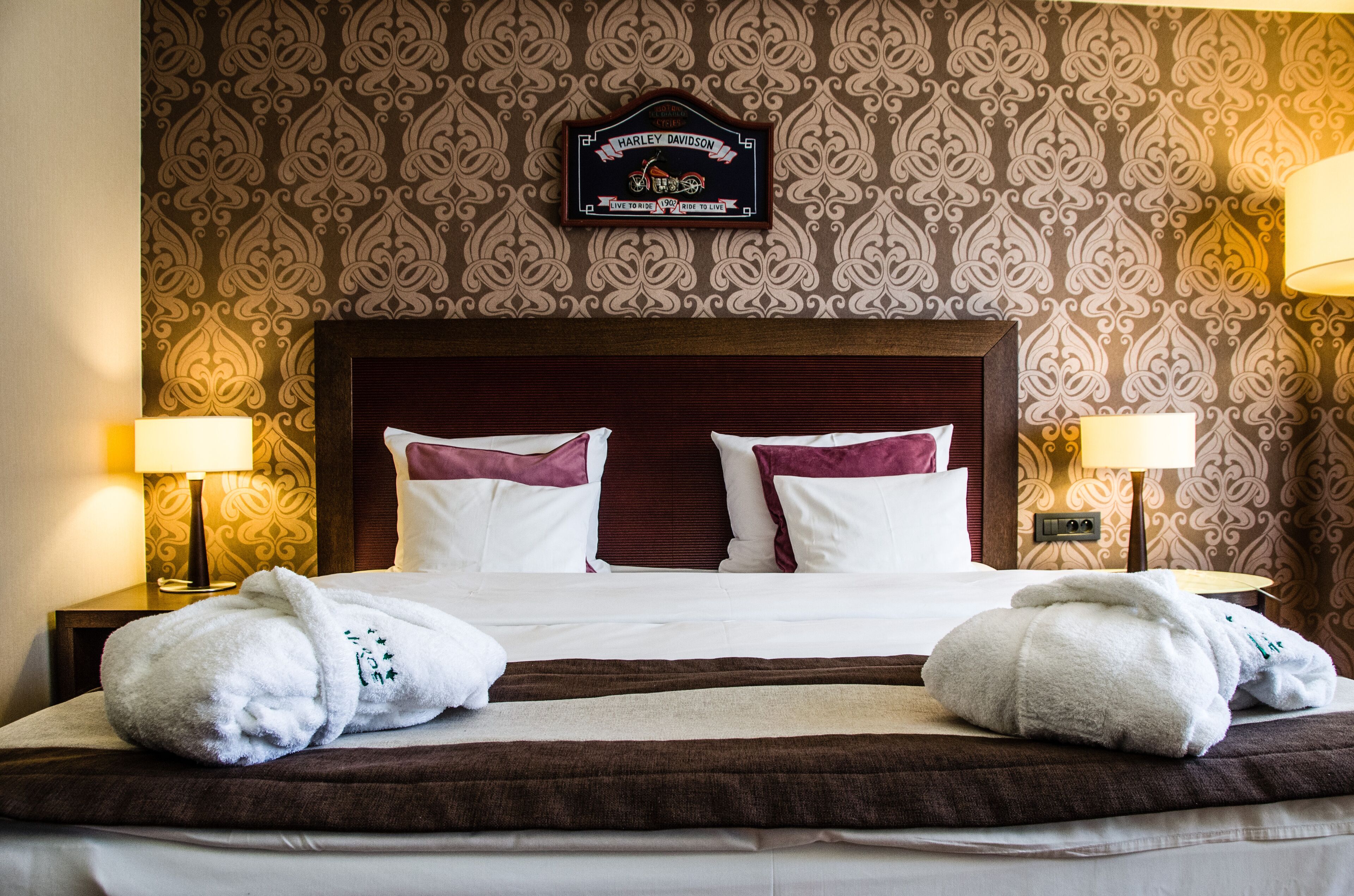 A hotel room with a patterned wallpaper, a Harley Davidson sign, a neatly made bed with white and maroon pillows, and two bedside lamps.