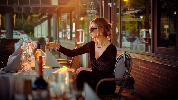 Una persona sentada en una mesa al aire libre con una copa de vino, un menú y una botella de vino. El lugar parece ser un restaurante o café con una pared de ladrillo y una ventana que muestra vista a la calle.