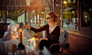Una persona sentada en una mesa al aire libre con una copa de vino, un menú y una botella de vino. El lugar parece ser un restaurante o café con una pared de ladrillo y una ventana que muestra vista a la calle.