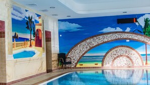 Una piscina interior con un mural de una escena de playa, un arco decorativo y una pantalla digital en el techo.